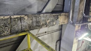 Viga com concreto desplacado e armadura exposta em ambiente industrial de caldeira, analisada em laudo técnico estrutural pela MT Engenharia