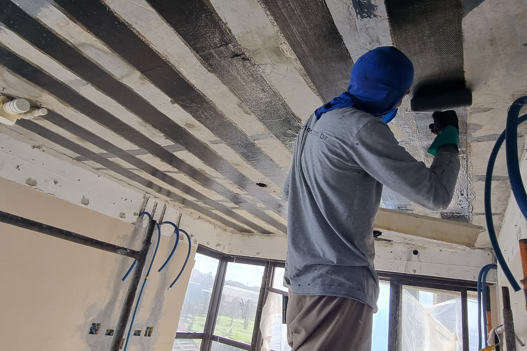 Operário aplicando manta de fibra de carbono com rolo para reforço estrutural em laje de concreto.